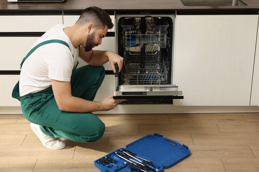 A technician kneeling at an open built-in dishwasher with a tool case on the kitchen floor