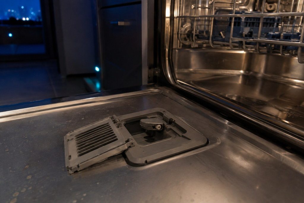 The drying vent in the inner face of an open dishwasher door with its cover unclipped and lifted aside