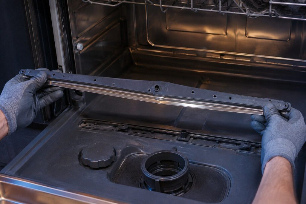 A flat wash bar lifted out of the floor of a dishwasher tub on two gloved hands
