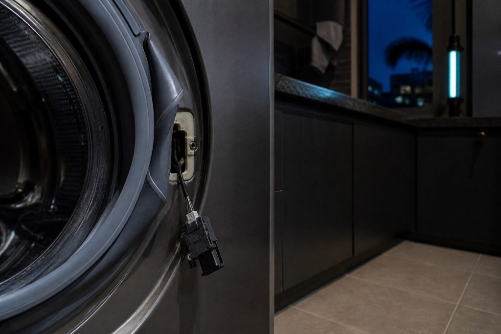 A door lock unit drawn out of the front panel of a washing machine and hanging on its wiring loom