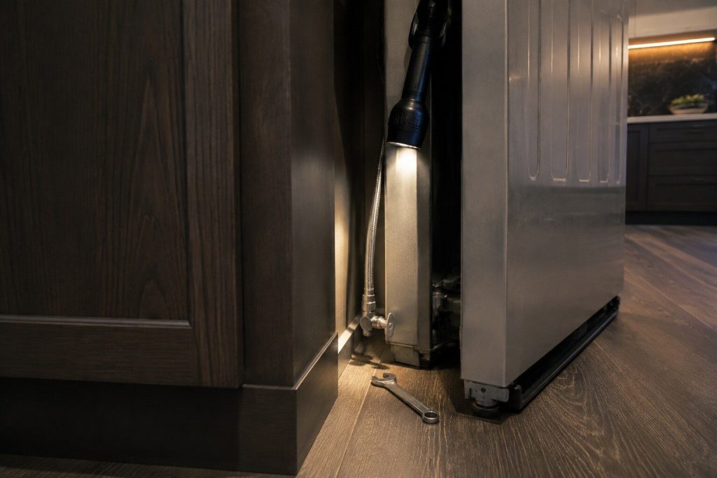 The braided water line and its small shut-off valve in the narrow gap behind a built-in refrigerator
